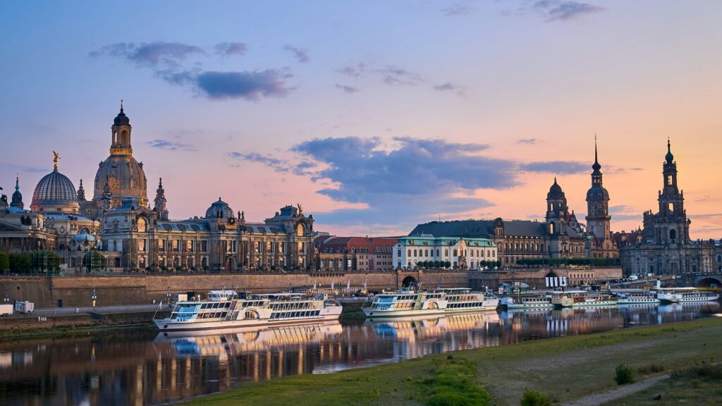 Abendliche Skyline von Dresden mit Frauenkirche, Elbufer und historischen Gebäuden, gesehen vom anderen Elbufer mit beleuchteten Schiffen.